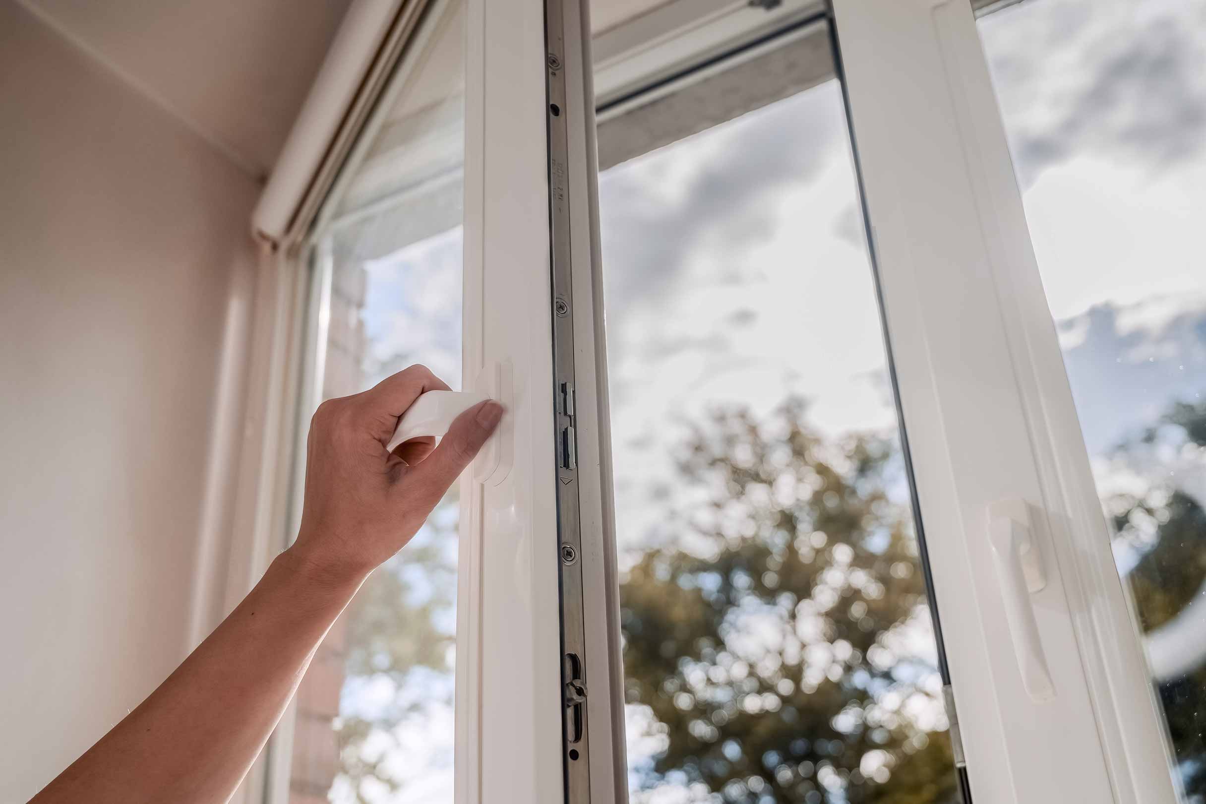 Eine Person öffnet ein Kunstofffenster an einem sonnigen Tag und lässt frische Luft in den Raum.